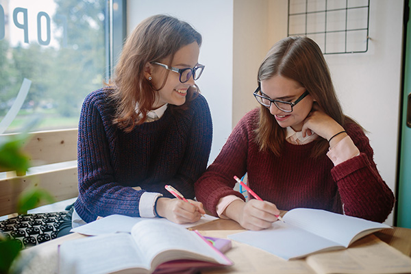 Two teenagers in glasses do homework together, adult girls laugh, they hold a pencil and pen in their hands to take notes in notebooks and textbooks
