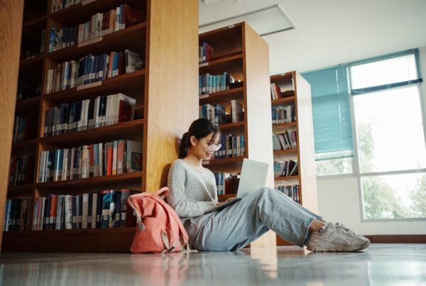 Student studying with laptop while sitting on the floor next to bookshelves in a library.